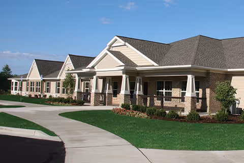 Exterior view of a single-story brick building with multiple peaked roofs, a covered porch with white columns, and well-maintained landscaping including green grass and small bushes under a clear blue sky.