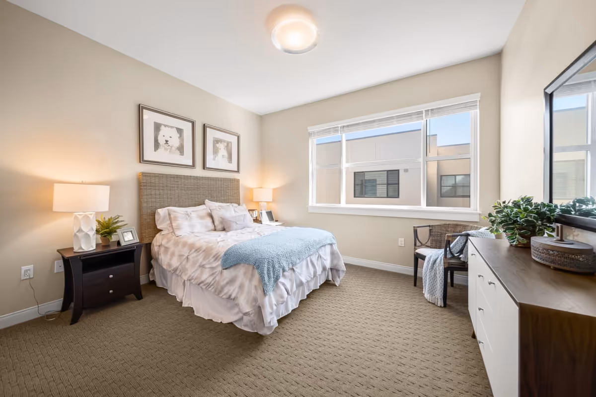 A bright and cozy bedroom with a large window letting in natural light. The room features a bed with a woven headboard, white and beige bedding, and a light blue throw blanket. On either side of the bed are dark wood nightstands with lamps and small decorative items. There are two framed pictures of dogs hanging above the bed. A wooden dresser with a plant and decorative items sits against the opposite wall, next to a chair with a blanket draped over it. The carpet is beige with a subtle pattern.
