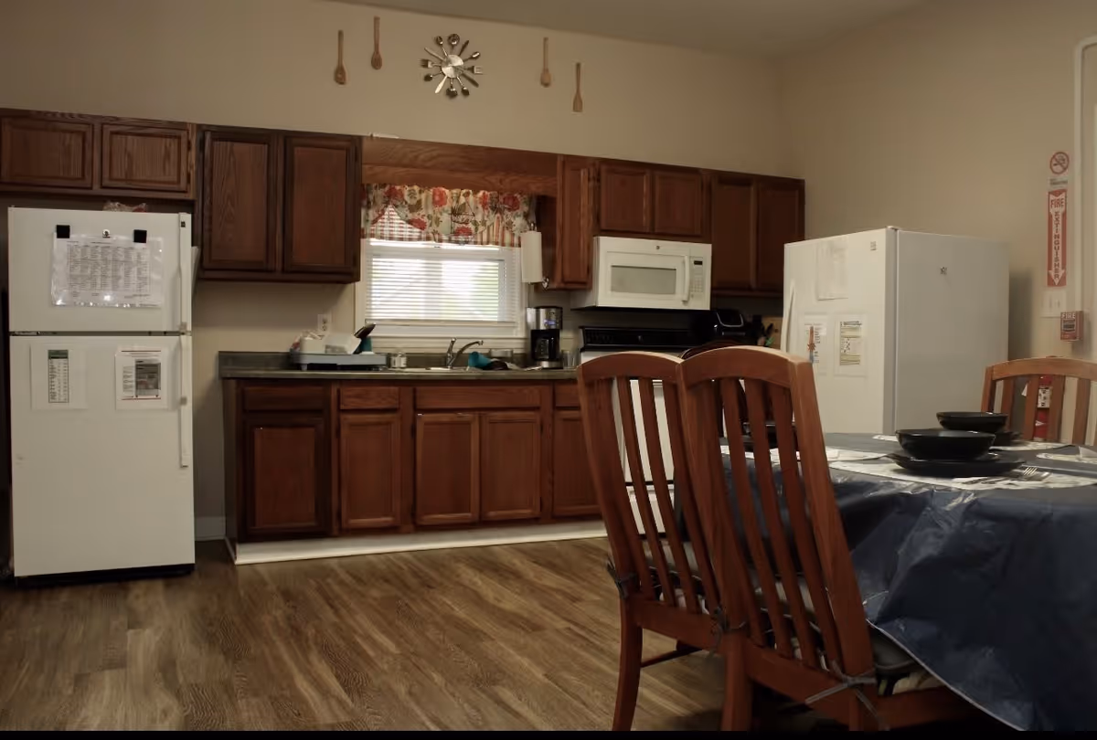 A kitchen area with wooden cabinets, a white refrigerator, a microwave above a stove, a coffee maker on the counter, and a window with floral curtains. In the foreground, there is a dining table covered with a dark tablecloth and wooden chairs around it. The floor is wood-patterned, and there are some kitchen utensils hanging on the wall above the window.