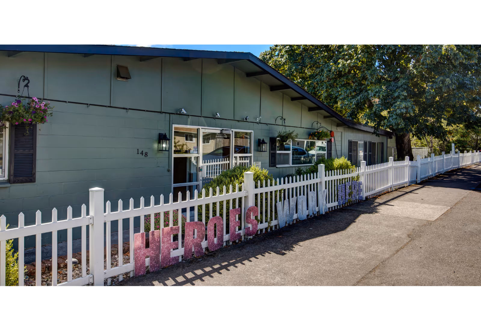 Front exterior of a single-story facility with a white picket fence, hanging flower baskets, and large decorative letters reading 'HEROES' along the fence.