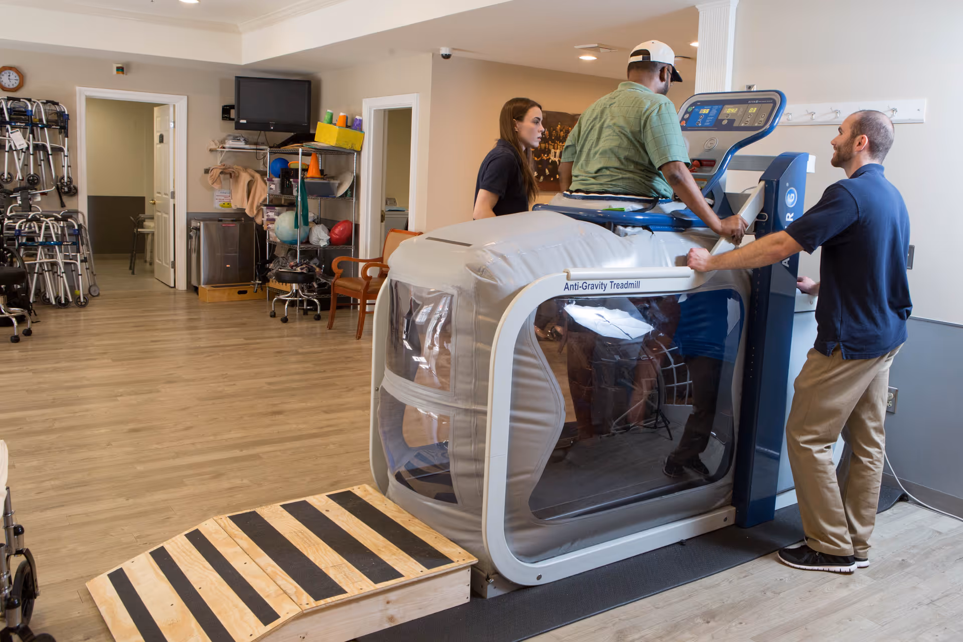 A man using an anti-gravity treadmill assisted by two staff members in a rehabilitation or therapy room with medical equipment and walkers visible in the background.
