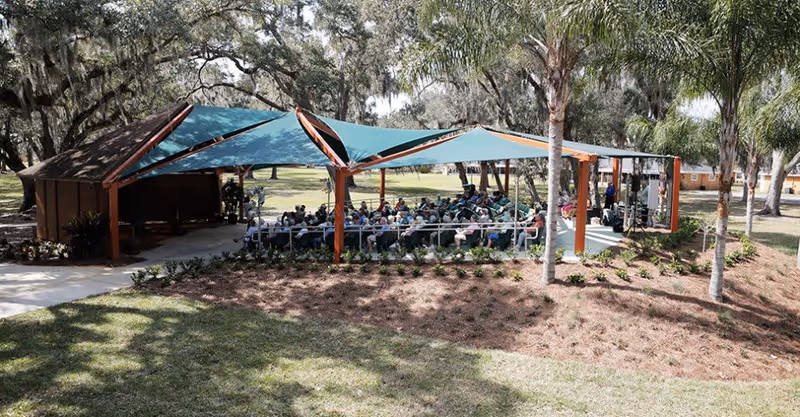 Outdoor pavilion with a large group of people seated under blue shade sails supported by wooden beams, surrounded by trees and landscaped grounds at Good Samaritan Society - Kissimmee Village - Village West.