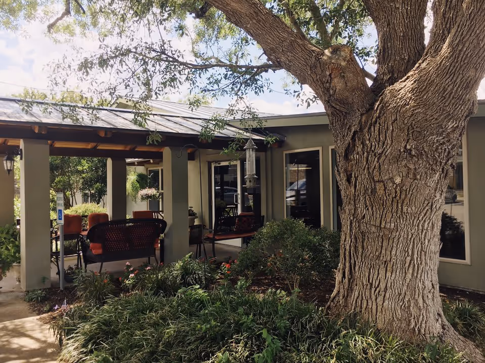 Outdoor seating area at Henry House Assisted Living LLC with a large tree in the foreground, surrounded by greenery and flowers. The seating area is covered with a roof and has several chairs and benches with orange cushions. The building exterior is visible with windows and doors.