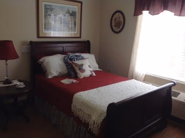 A cozy bedroom with a dark wooden bed frame, a red bedspread, white and decorative pillows, a white throw blanket, a nightstand with a red lamp and a cup, a framed picture above the bed, and a window with sheer curtains and a dark valance.