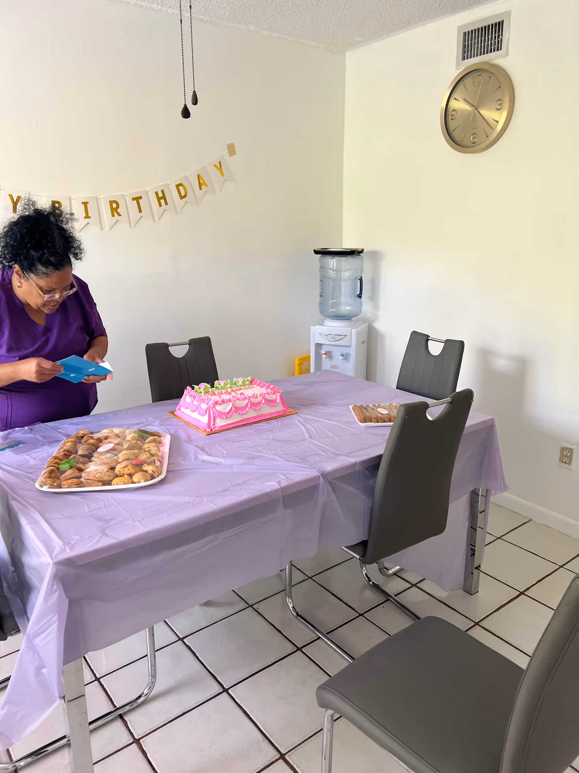 A woman in a purple outfit standing next to a dining table covered with a lavender tablecloth. On the table, there is a pink birthday cake and trays of assorted cookies. A 'Happy Birthday' banner is partially visible on the wall behind her. The room has white tiled floors, gray chairs, a wall clock, and a water dispenser in the corner.