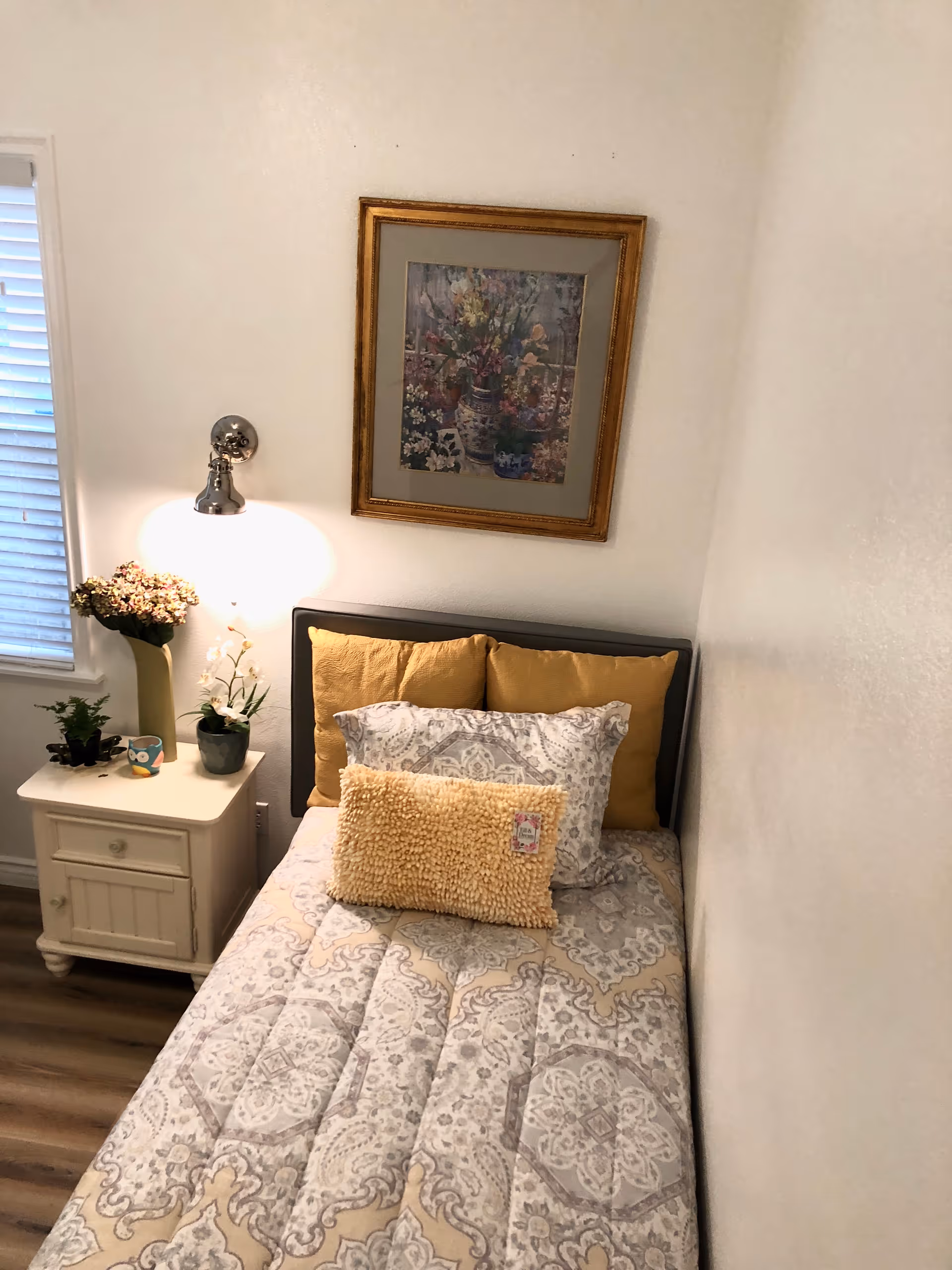 A small bedroom with a single bed against the wall. The bed has a patterned comforter in shades of gray and beige, with three mustard yellow pillows and one textured yellow pillow. Next to the bed is a white nightstand with a drawer and cabinet, holding three potted plants and a small decorative item. Above the bed hangs a framed floral painting with a gold frame. A wall-mounted lamp provides light, and a window with white blinds is partially visible on the left.