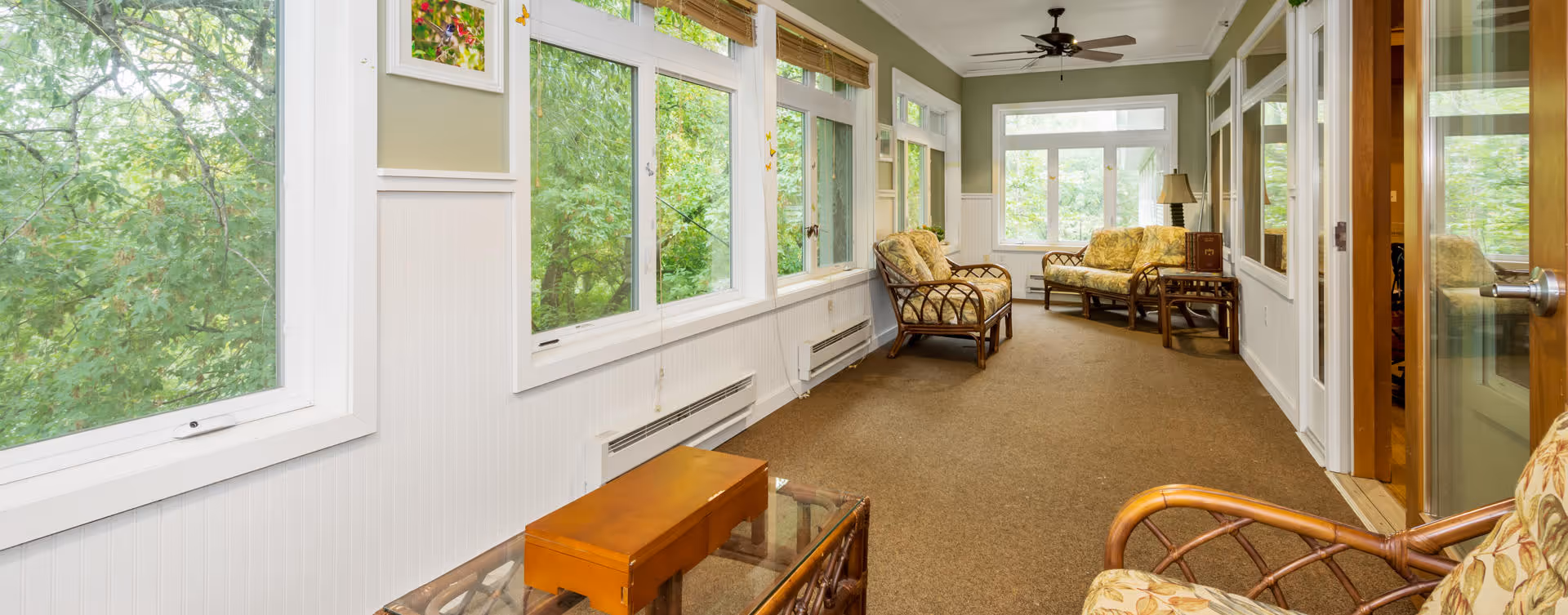 A bright and cozy sunroom with large windows overlooking green trees. The room features wicker furniture with floral cushions, including chairs and a loveseat, a glass-top coffee table, and a ceiling fan. The walls are painted green above white wainscoting, and the floor is carpeted in brown.