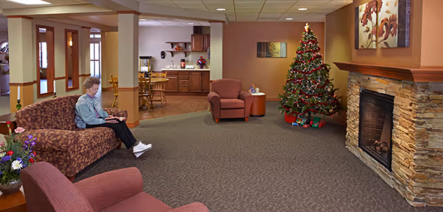 A cozy senior living facility common area with a decorated Christmas tree, a stone fireplace, and comfortable seating including a patterned sofa and armchairs. An elderly woman is sitting on the sofa reading a book. The background shows a kitchen area with wooden cabinets and a dining table with chairs.