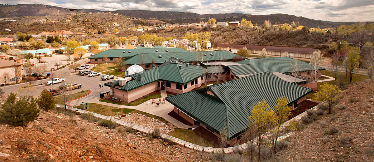 Aerial view of Sunshine Gardens West Assisted Living facility showing multiple connected buildings with green roofs surrounded by trees and a parking lot, set against a backdrop of hills and a partly cloudy sky.
