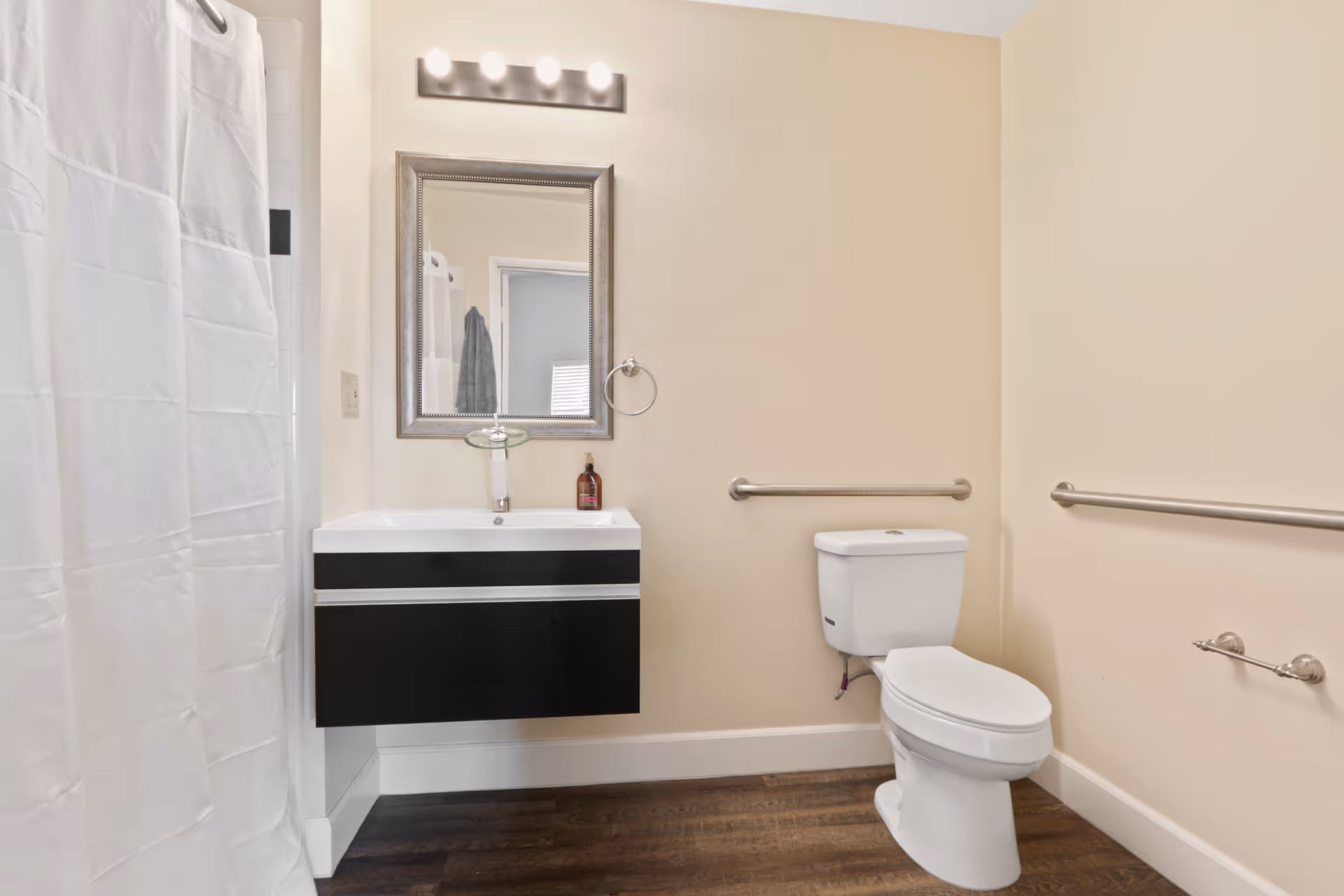 A clean bathroom with beige walls and dark wood flooring. It features a white toilet with grab bars on both sides, a modern black and white floating sink vanity with a mirror above it, and a shower with a white curtain on the left side.