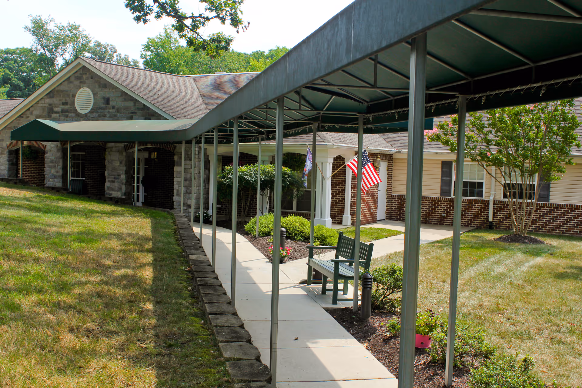 Covered walkway leading to the entrance of a brick-and-siding senior living building with a bench and American flags.