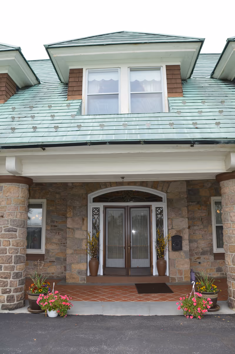 Entrance of a building with a stone facade, double glass doors with decorative side panels, a green roof with dormer windows, and flower pots with colorful flowers on either side of the entrance.