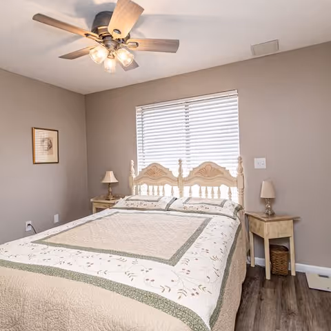 A bedroom with a double bed featuring a decorative quilt and two pillows. The bed has a carved wooden headboard. On each side of the bed, there are wooden nightstands with lamps. A ceiling fan with lights is mounted on the ceiling. A window with closed blinds is behind the bed, and a framed picture hangs on the left wall. The floor is wood, and the walls are painted beige.