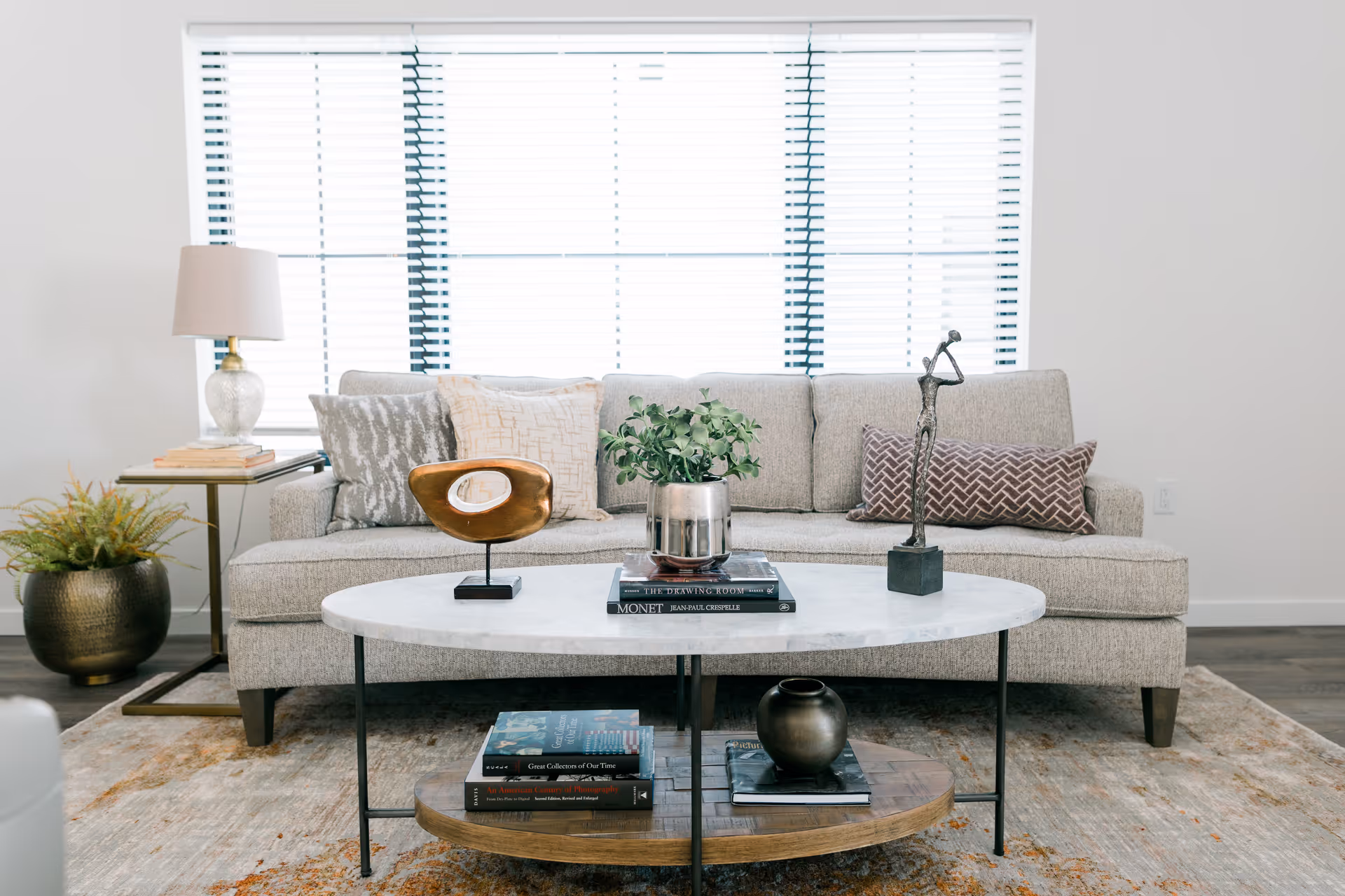 Bright living room with a beige sofa, patterned pillows, a round coffee table with books and decor, and a large window with blinds behind it.