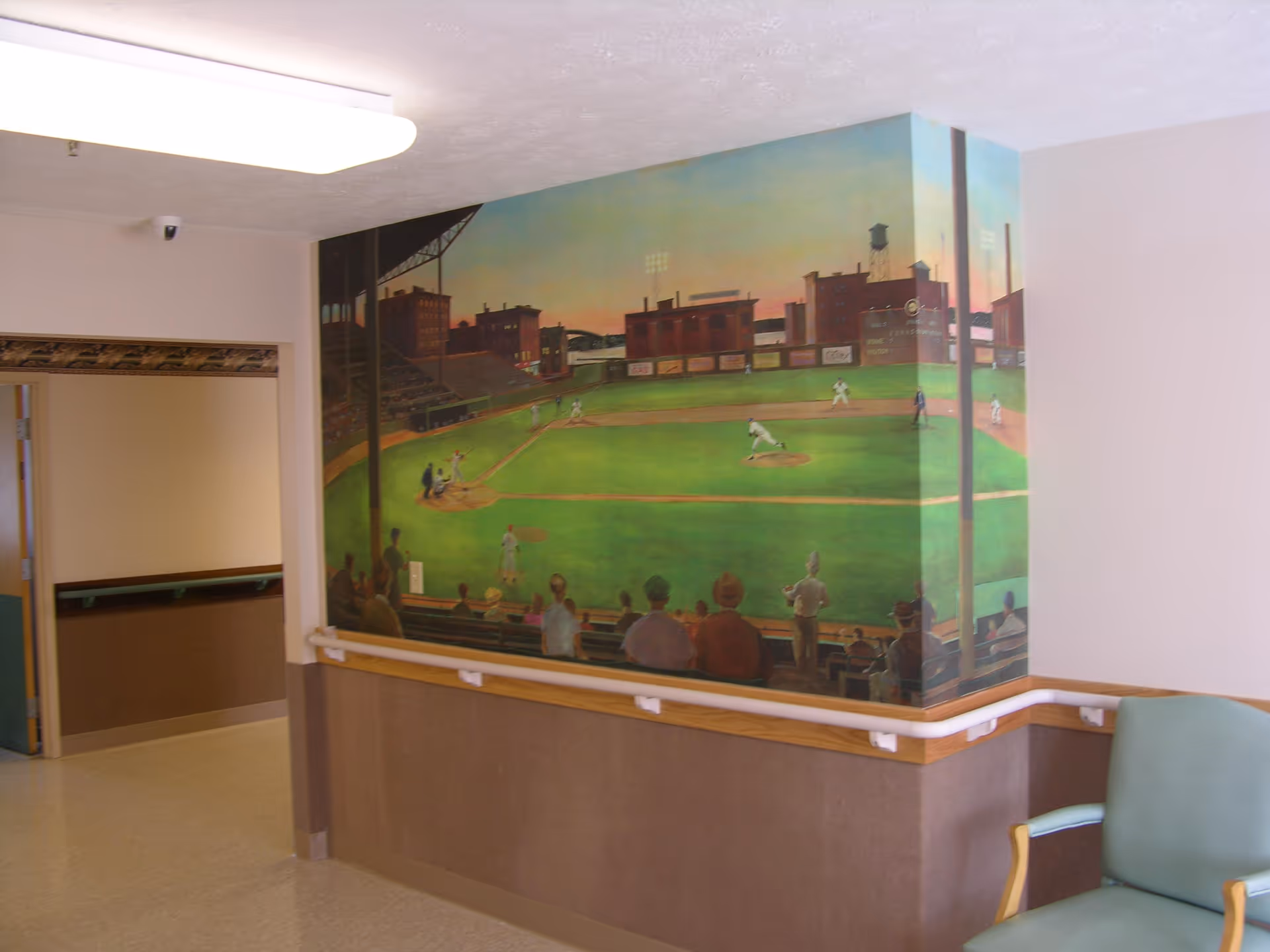 Interior hallway corner of a senior living facility with a mural depicting a baseball game on the wall. The hallway has handrails along the walls and a green cushioned chair is partially visible on the right side.