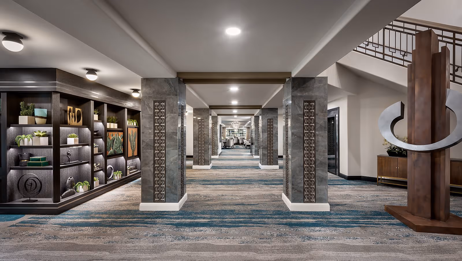 A spacious, well-lit interior hallway with decorative columns and patterned carpet. On the left side, there is a built-in dark wood shelving unit displaying various decorative items and plants. On the right side, there is a large modern sculpture near a staircase with a metal railing. The hallway extends into a seating area with chairs and a chandelier in the distance.