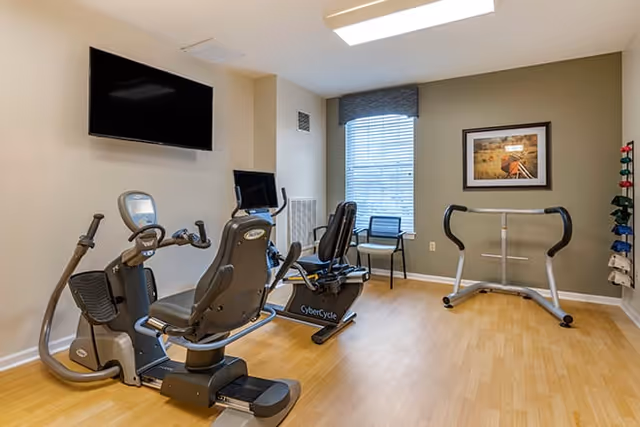 A small exercise room with two recumbent stationary bikes, a TV mounted on the wall, a window with blinds, a chair, a framed picture on the wall, and a rack holding colorful dumbbells.