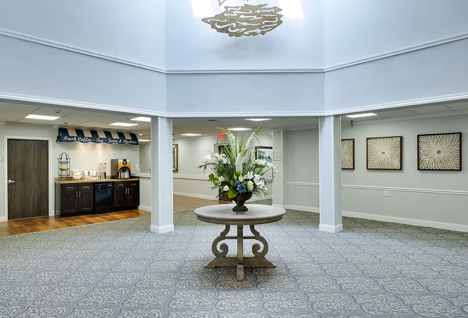 A spacious interior area with a round wooden table in the center holding a large floral arrangement. The room has patterned carpet flooring and white walls with decorative molding. On the left side, there is a small refreshment area with a sign above it that reads 'Fresh Coffee - Tea - Juice & Pastries' and a counter with coffee and beverage dispensers. The ceiling is high with a skylight and a decorative light fixture. Three framed artworks are visible on the right wall.