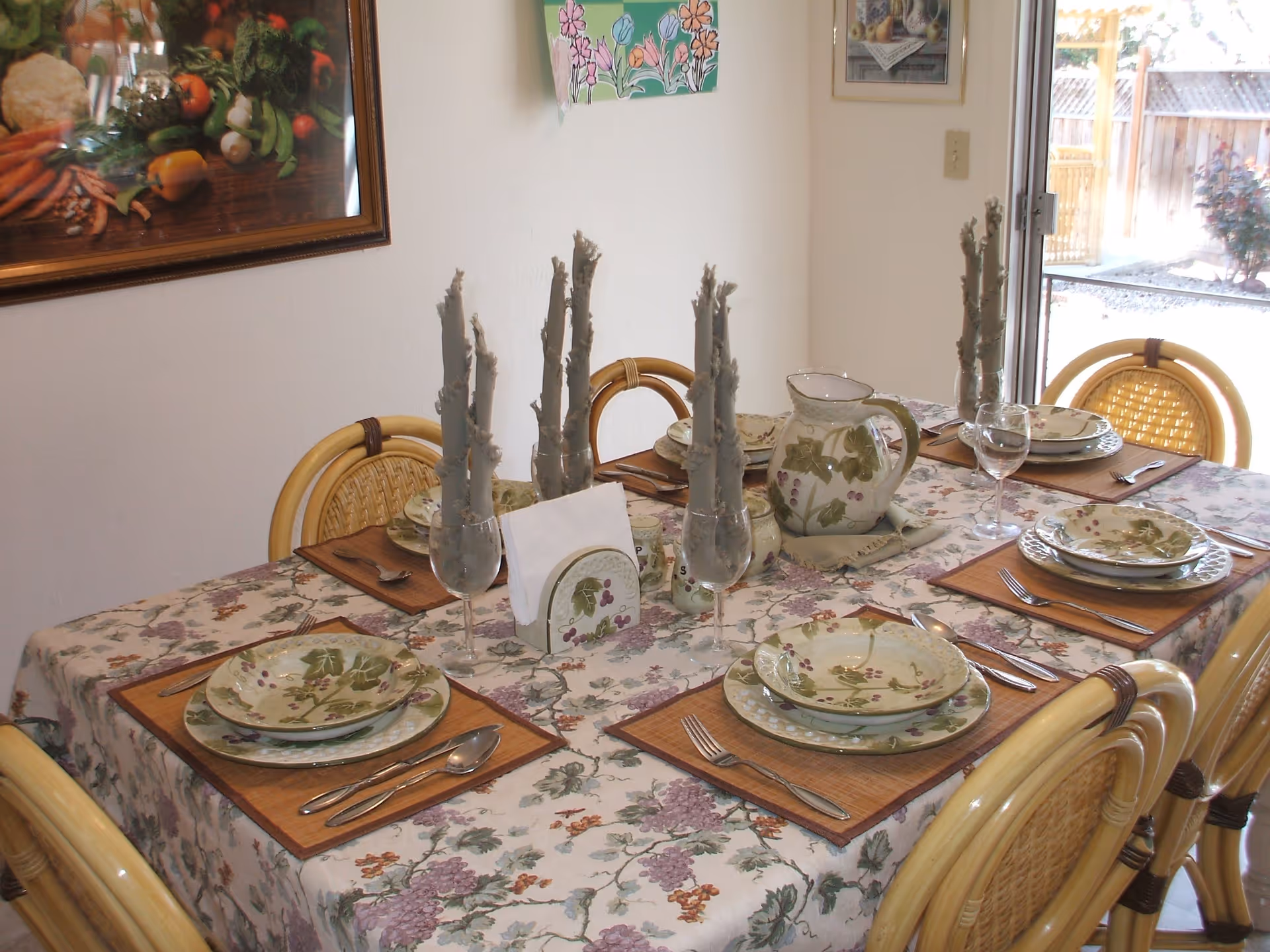 A dining table set for six with floral dishes, placemats, candles and a pitcher in a bright dining room.