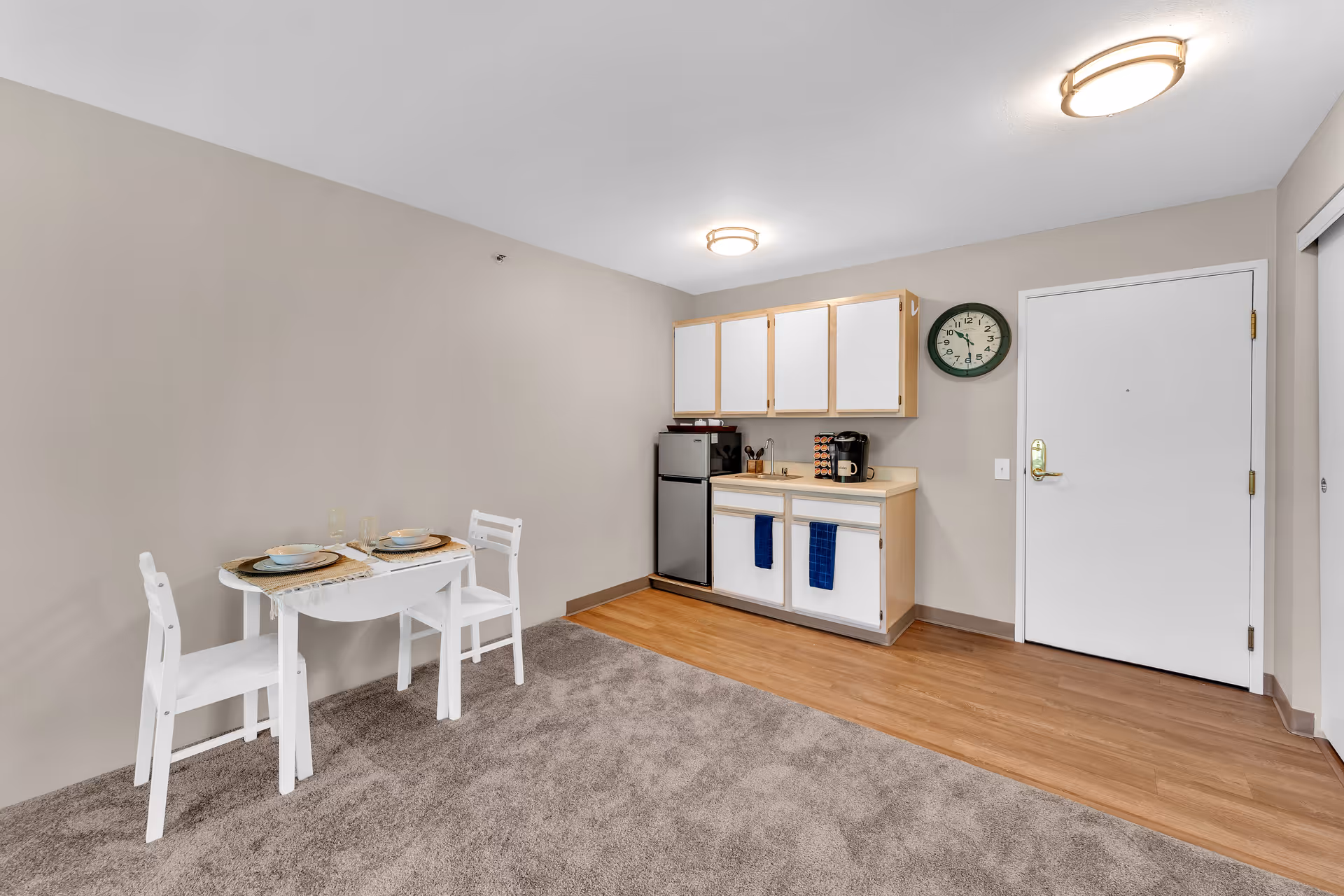 Small kitchenette area with white cabinets, a mini refrigerator, a coffee maker, and a sink. There is a round wall clock above the counter and a white door to the right. A small white dining table with two chairs is set with plates and glasses on the left side. The floor is a combination of carpet and wood, and the walls are painted beige.