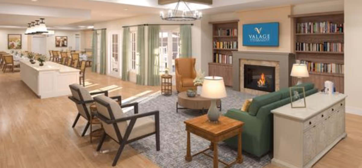 Bright open senior living common area with seating around a fireplace and bookshelves, adjacent to a dining/kitchen island.