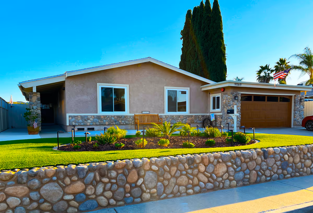 Front exterior view of a single-story residential building with a stone facade and beige walls. There is a well-maintained garden with green grass, small plants, and a wooden bench in front of the house. The driveway leads to a brown garage door, and an American flag is mounted near the garage. Tall trees and palm trees are visible in the background under a clear blue sky.