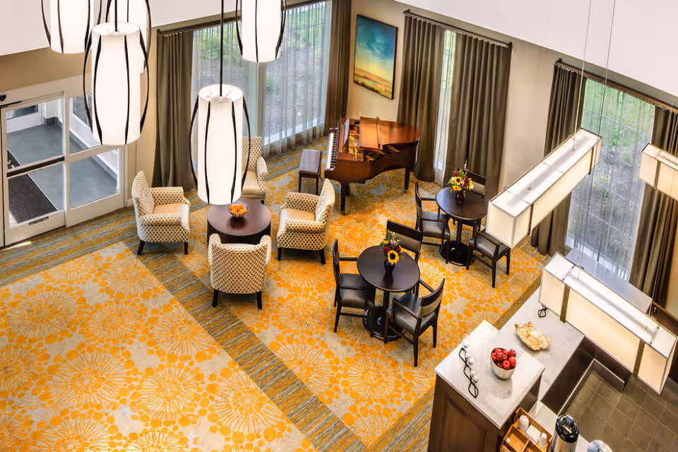 A bright and spacious common area with large windows covered by curtains, featuring a grand piano, round tables with chairs, armchairs arranged around a small table, and modern hanging light fixtures. The floor is covered with a patterned carpet in shades of yellow and beige, and there are decorative flowers on the tables.