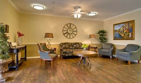 Comfortable common living area with a sofa, armchairs, round coffee table, wall clock, lamps, and artwork on the walls.