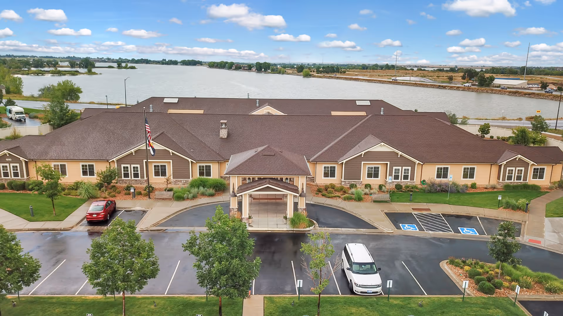 Aerial view of a single-story senior living facility named Seven Lakes Memory Care, with a brown roof and beige exterior walls. The building is surrounded by a parking lot with several cars, including two handicap parking spaces near the entrance. There are landscaped areas with trees and shrubs around the building. In the background, there is a large body of water and a partly cloudy sky.