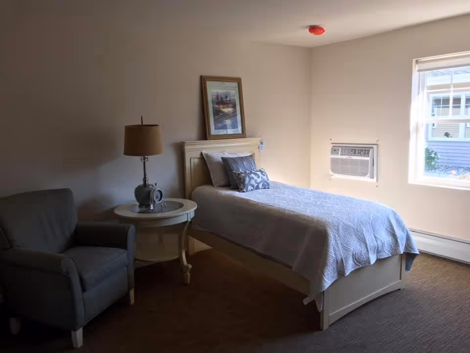 A simple bedroom with a single bed covered in a light gray quilt and decorative pillows. Next to the bed is a round white side table with a lamp and a clock. There is a gray armchair to the left of the table. A framed picture is hung on the wall above the bed's headboard. A window with an air conditioning unit is on the right wall, allowing natural light into the room.
