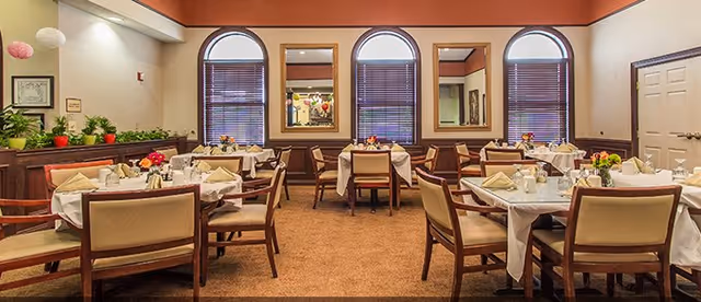 A dining room with multiple tables covered in white tablecloths, each set with napkins, glasses, and small floral centerpieces. The room features three large arched windows with blinds, mirrors on the wall between the windows, and a warm color scheme with beige walls and brown carpet. There are wooden chairs with beige cushions around the tables and some green plants on a ledge along the left wall.