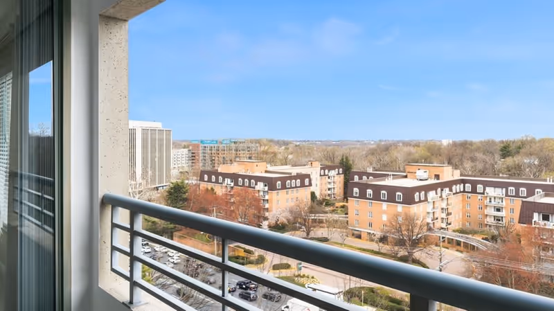 View from a balcony overlooking a residential complex with multiple buildings, trees, and a street with cars. The sky is clear and blue.