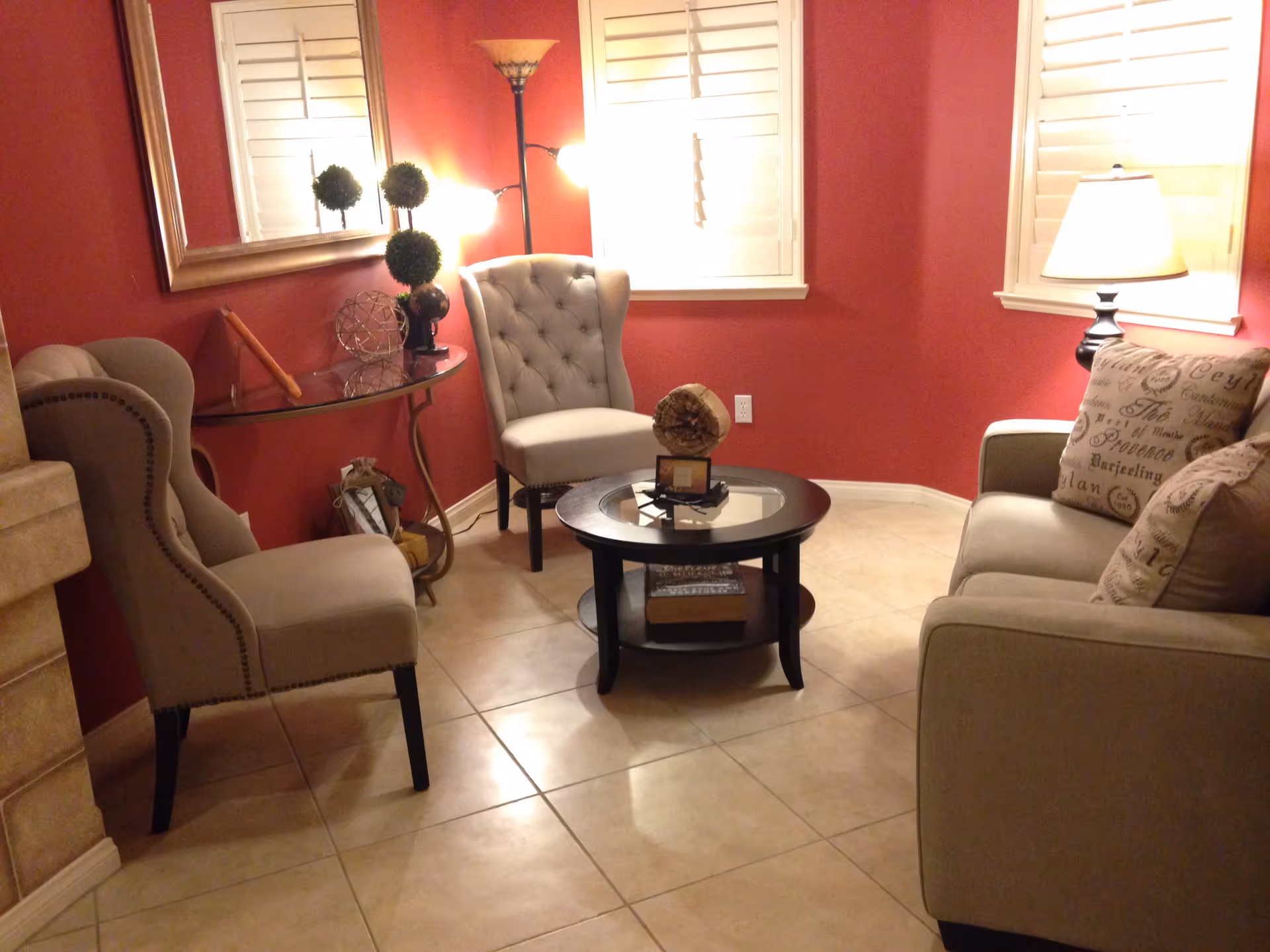 A cozy living room with red walls, beige tiled floor, and white window shutters. The room features a beige sofa with patterned pillows, two beige tufted armchairs, a round glass-top coffee table with decorative items, a glass console table with decor, a floor lamp, and a table lamp providing warm lighting.