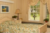 A cozy bedroom with a single bed covered in a floral patterned bedspread. Next to the bed is a nightstand with a lamp and a small plant. The room has a large window with decorative curtains, allowing natural light to brighten the space. There is a framed picture on the wall above the bed.