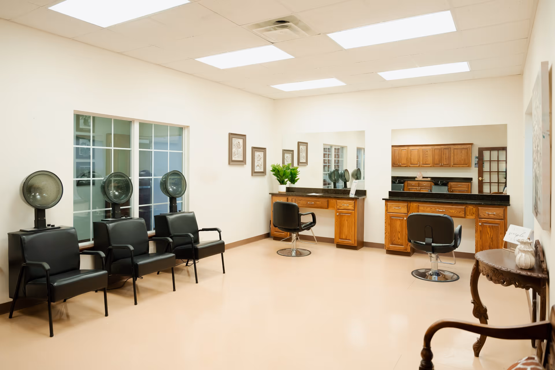 Interior view of a hair salon area in a senior living facility with three black salon chairs under hair dryers along one wall, two styling stations with mirrors and black chairs, wooden cabinets, and a small decorative table with a chair in the foreground.