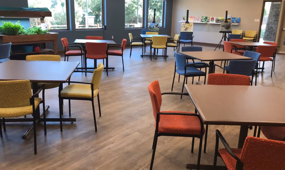 Spacious common room with multiple tables and colorful chairs and large windows.