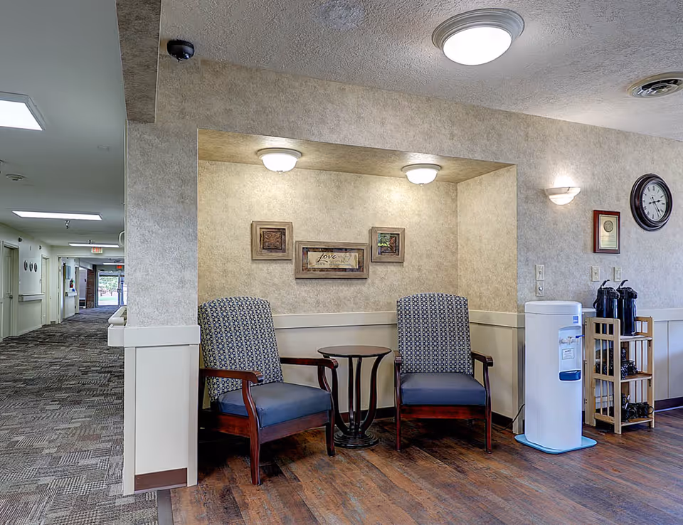 A cozy seating area in a hallway with two patterned armchairs and a small round table between them. The walls are beige with framed artwork and soft lighting fixtures above the chairs. To the right, there is a water cooler and a wooden rack holding coffee pots and supplies. The hallway extends to the left with carpeted flooring and an exit door visible in the distance.