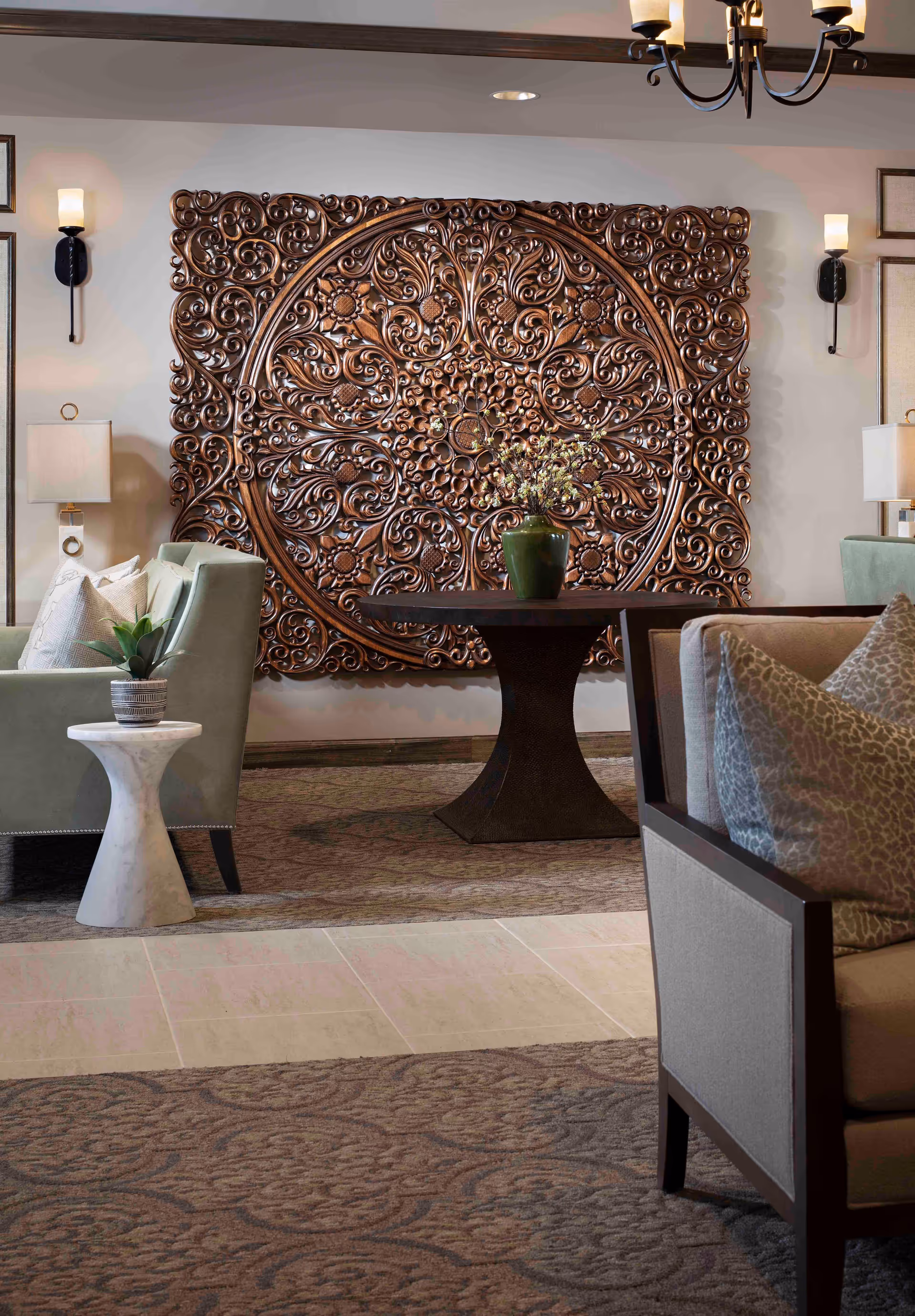 A cozy senior living room area featuring a large, ornate wooden wall decoration with intricate floral and scrollwork patterns. The room includes comfortable armchairs with cushions, a round wooden table with a green vase holding flowers, a small marble side table with a potted plant, wall sconces, and soft lighting creating a warm ambiance.
