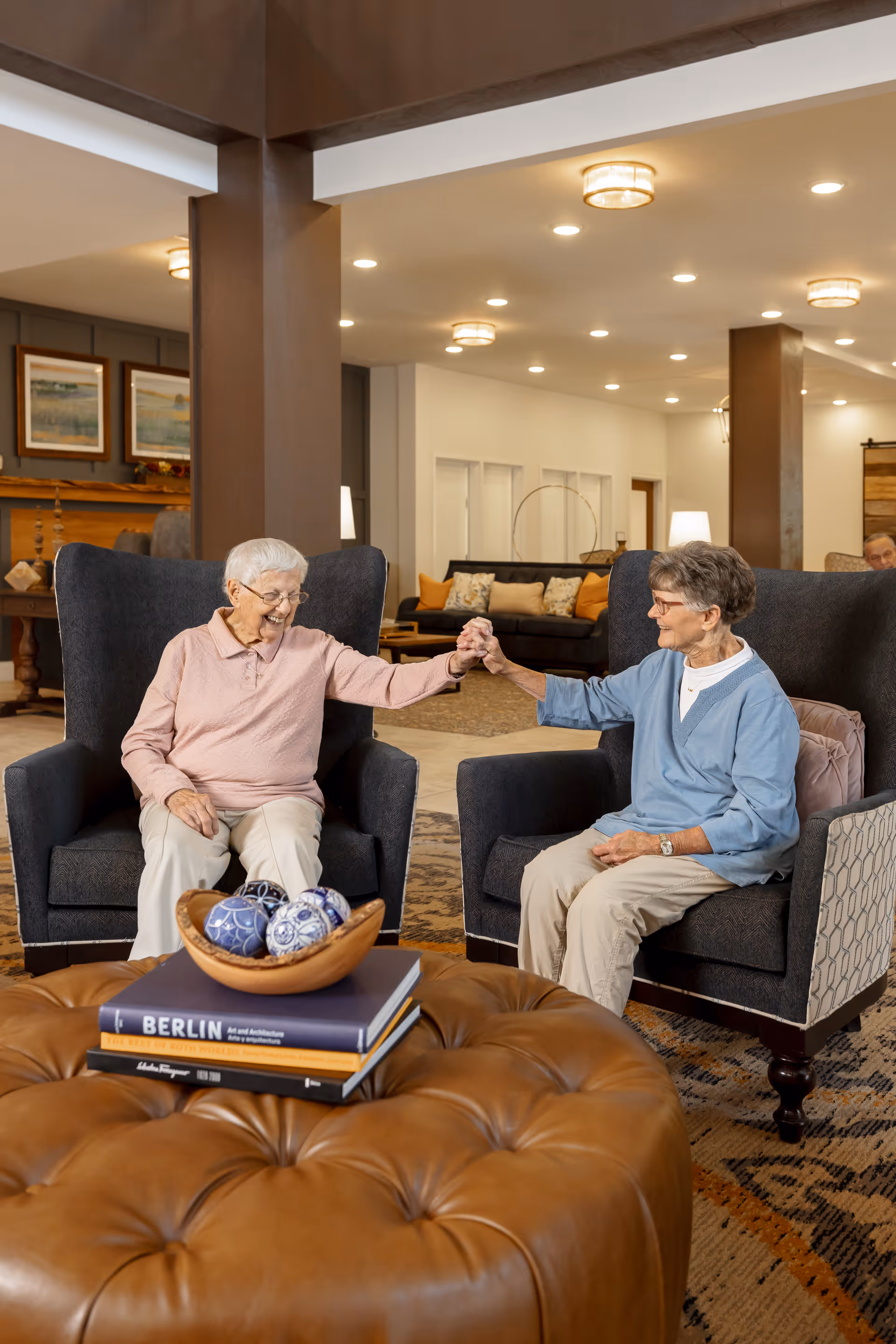 Two elderly women sitting in dark armchairs in a well-lit living room area, holding hands and smiling at each other. In front of them is a large brown tufted ottoman with a wooden bowl containing decorative balls and a stack of books. The background shows a cozy seating area with couches, pillows, and framed artwork on the walls.