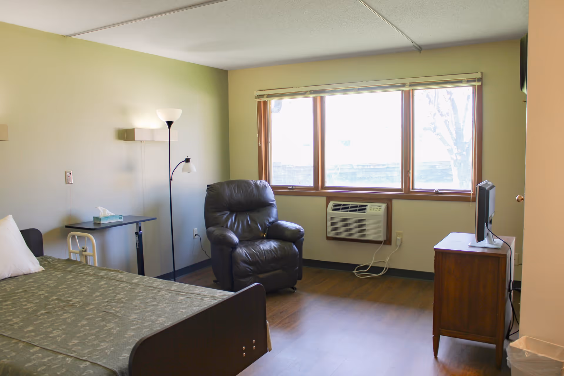 A sparsely furnished senior living bedroom with a bed, leather recliner, small TV on a dresser, and a large window with a wall air conditioner.