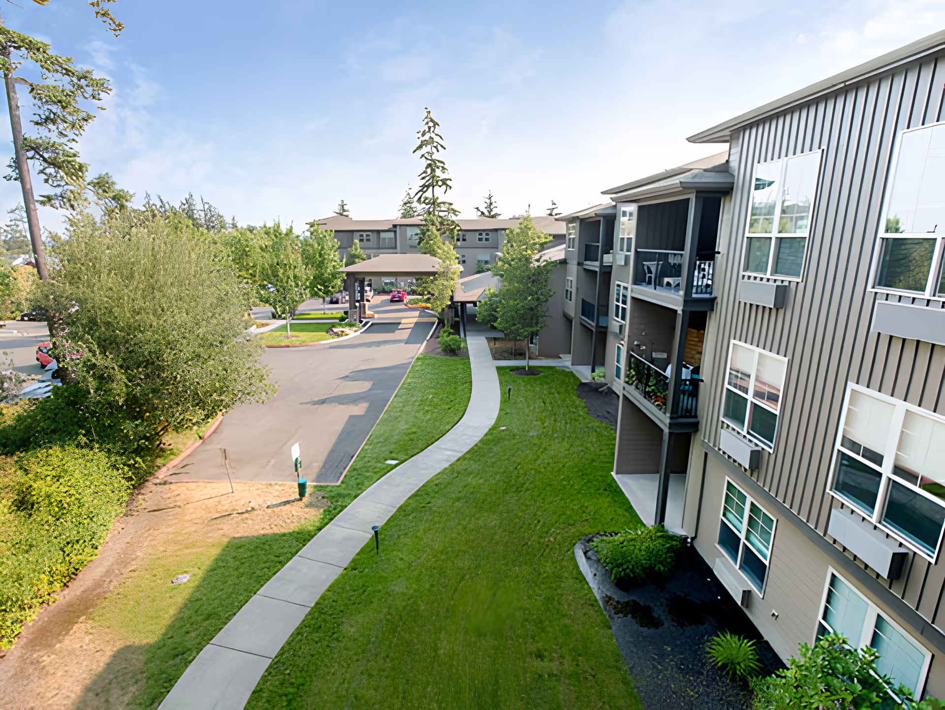 Exterior view of a senior living facility with a paved driveway, green lawn, trees, and a multi-story building with balconies and large windows under a blue sky.