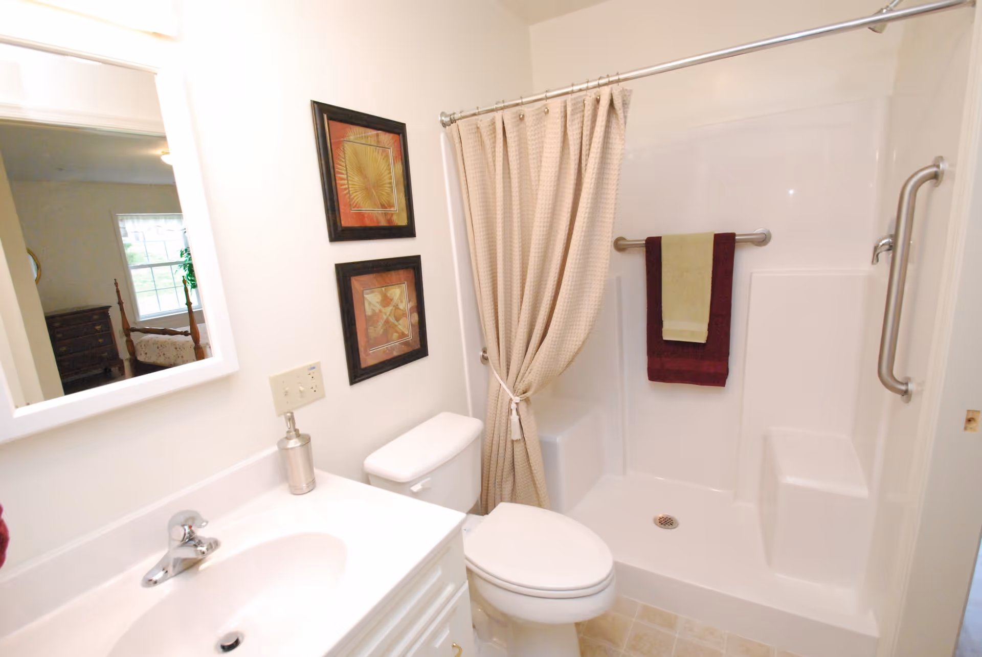 A clean bathroom featuring a white sink with a silver faucet, a toilet, and a shower area with a beige curtain tied to one side. Two framed artworks hang on the wall above the toilet, and two towels, one green and one burgundy, hang on a silver towel bar inside the shower. A mirror above the sink reflects part of a bedroom with a window and wooden furniture.
