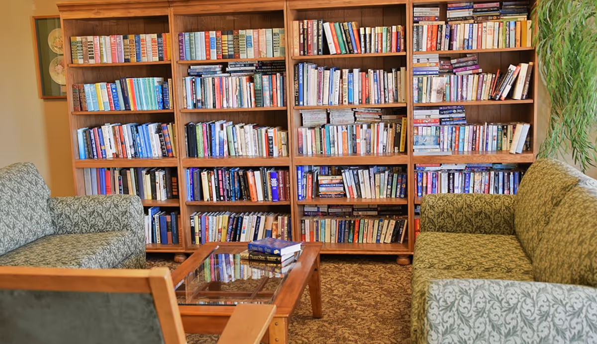 A cozy reading area with two patterned green sofas facing each other and a wooden coffee table with books on it. Behind the sofas is a large wooden bookshelf filled with many books. A green plant is visible on the right side of the image.