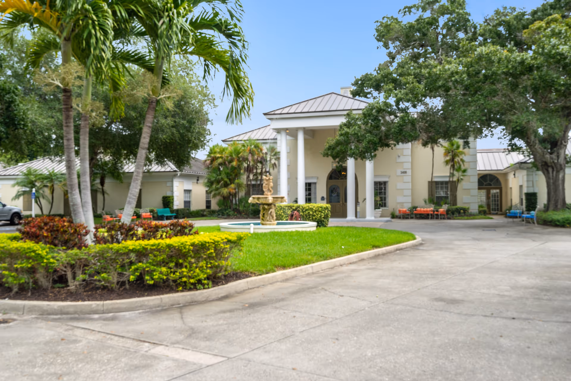 Front exterior view of Summerfield Senior Living facility showing a circular driveway with a central fountain, surrounded by green grass, palm trees, and other landscaping. The building has a light-colored facade with white columns at the entrance and several benches placed outside.