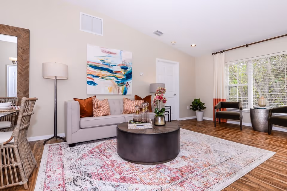 A bright living room with a light gray sofa adorned with orange and striped pillows, a round dark coffee table with decorative items, a colorful abstract painting on the wall, a tall floor lamp, two dark chairs near a large window with white blinds and curtains, and a patterned area rug on wooden flooring.