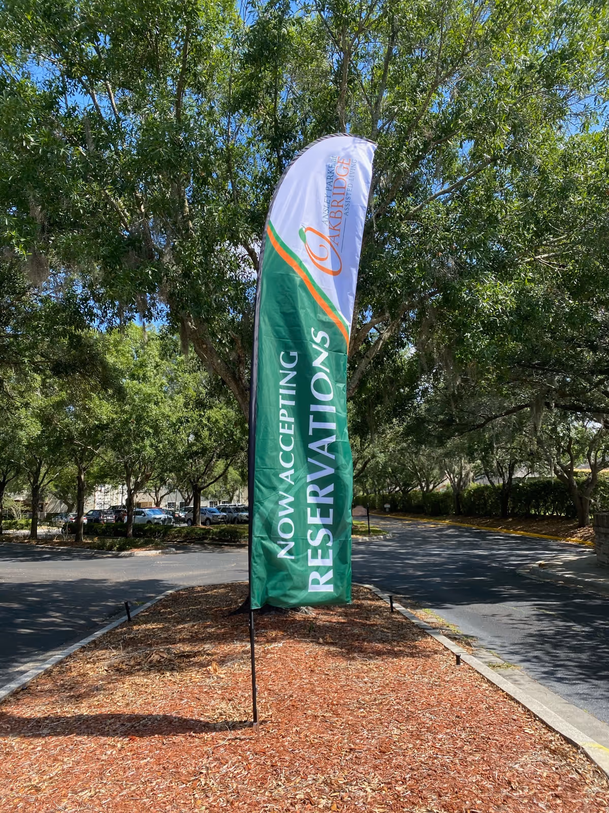 A tall vertical banner flag outdoors on a landscaped median strip with mulch and surrounded by trees and a parking lot. The banner reads 'NOW ACCEPTING RESERVATIONS' and has the Ansley Parke at Oakbridge logo at the top.