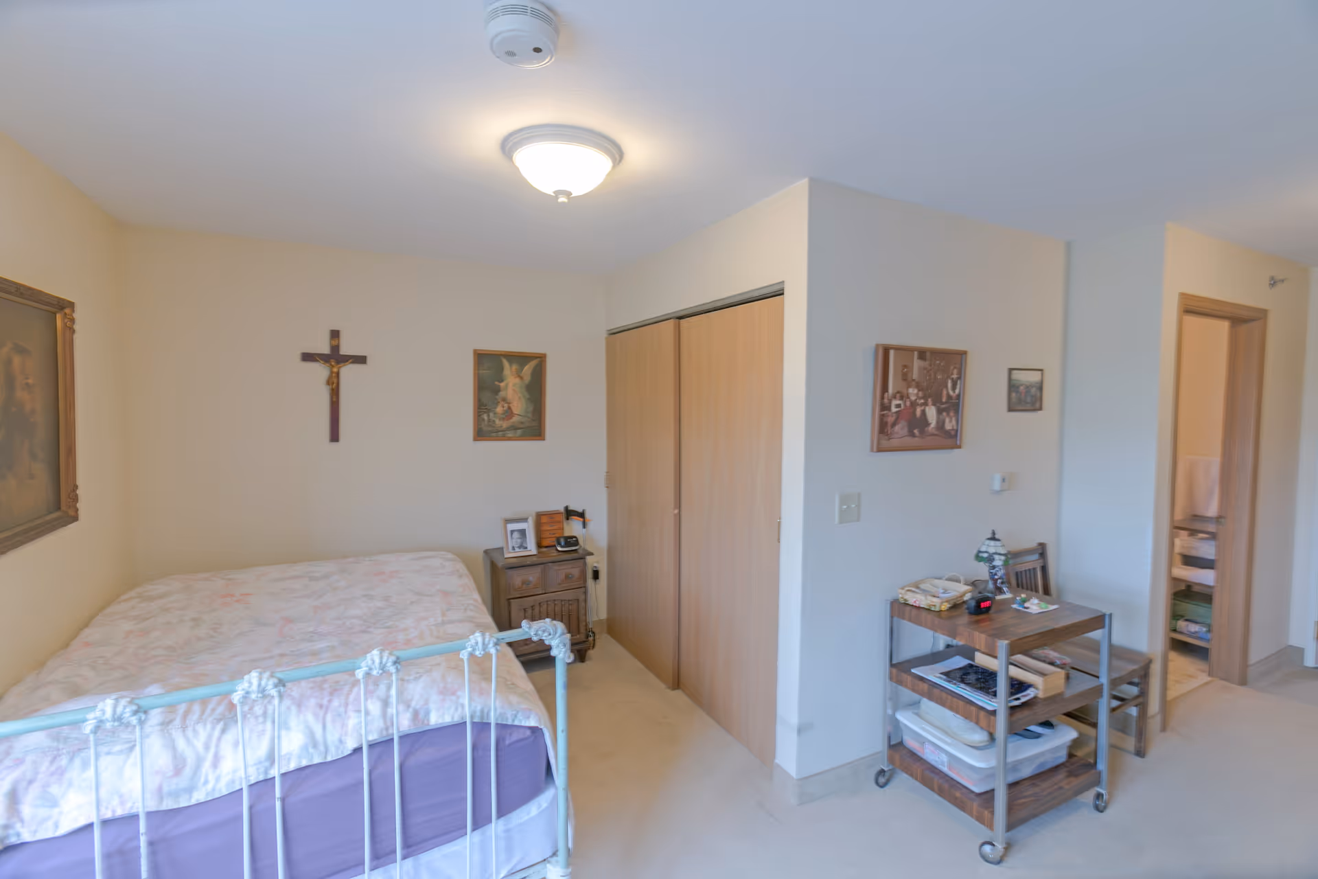 A bedroom in a senior living facility with a bed featuring a white metal frame and floral bedding. The room has beige walls adorned with religious artwork including a crucifix and framed pictures. There is a wooden nightstand next to the bed with personal items on it. A wooden closet with sliding doors is visible, along with a small wooden cart holding various items and a chair. An open doorway leads to another room with shelves and towels.