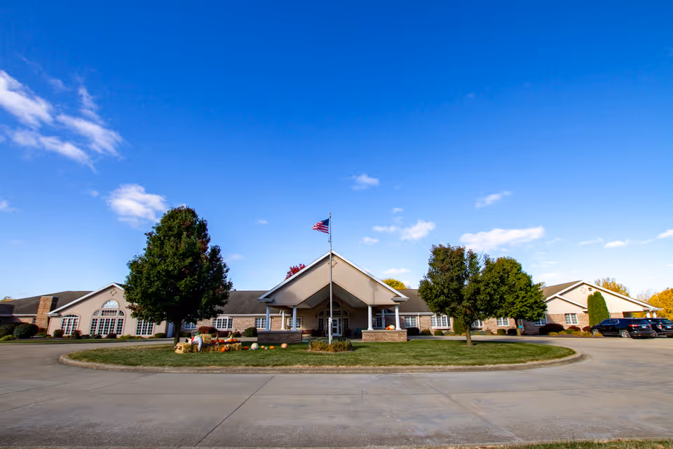 Front exterior view of Hickory Estates of Pana, a single-story building with a central entrance featuring a covered porch and an American flag on a flagpole. The building is surrounded by a circular driveway, green lawn, trees, and a few parked cars under a clear blue sky.