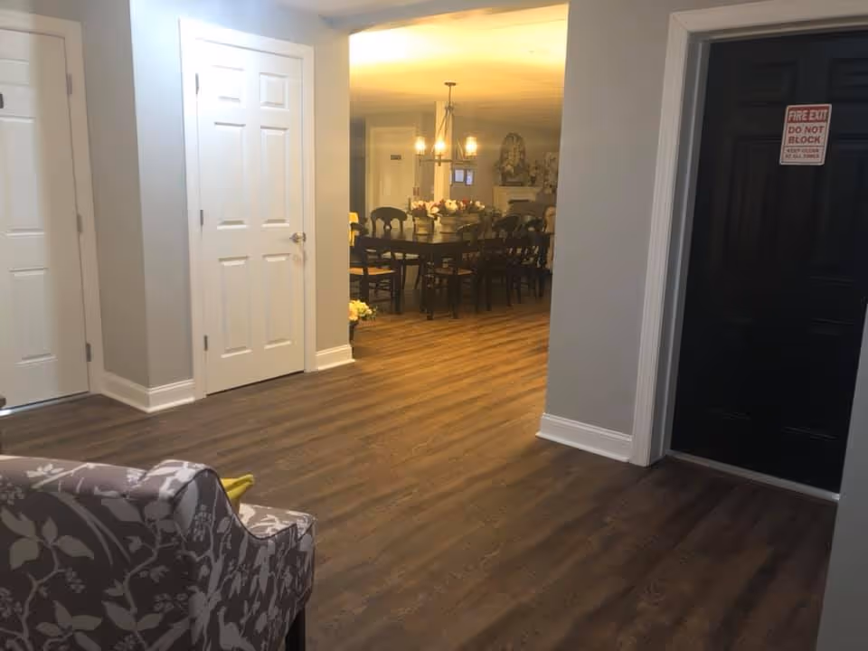 Interior view of a senior living facility showing a hallway with wood flooring, a floral patterned armchair in the foreground, white doors on the left, and a black door with a fire exit sign on the right. In the background, there is a dining area with a large table, chairs, a chandelier, and decorative items on the table and walls.