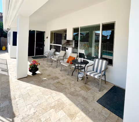Outdoor covered patio area with tiled flooring, featuring several chairs including striped and cushioned ones, a small round table, a potted plant with flowers, and large windows with blinds looking into the building.